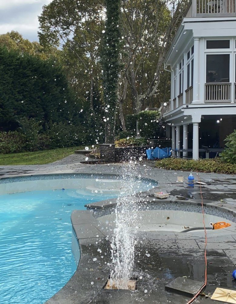 A fountain shoots a vertical jet of water into the air next to a swimming pool and spa on a patio outside a house.