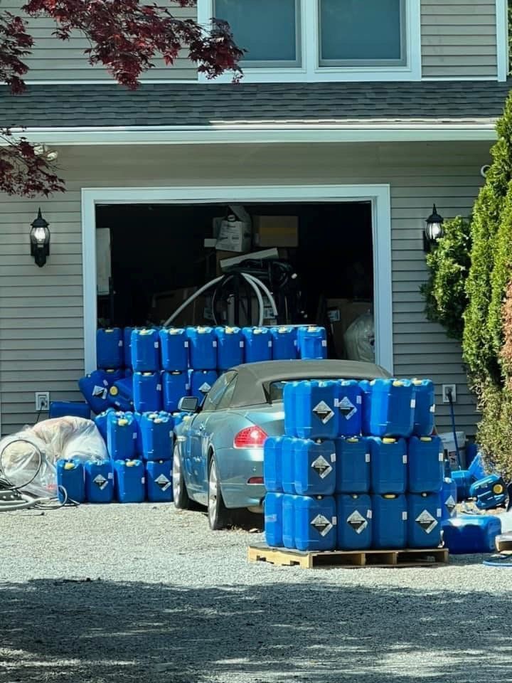 A blue convertible parked in a driveway in front of a garage stacked with many blue plastic chemical containers.