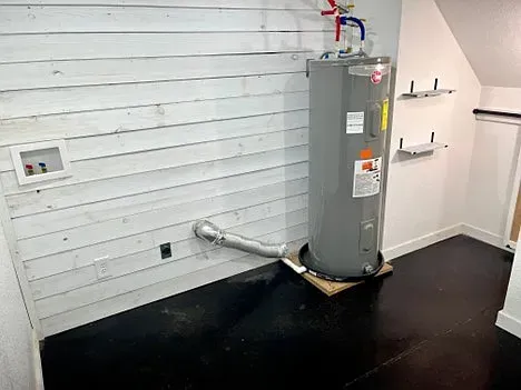 A water heater is sitting in a laundry room next to a white wall.