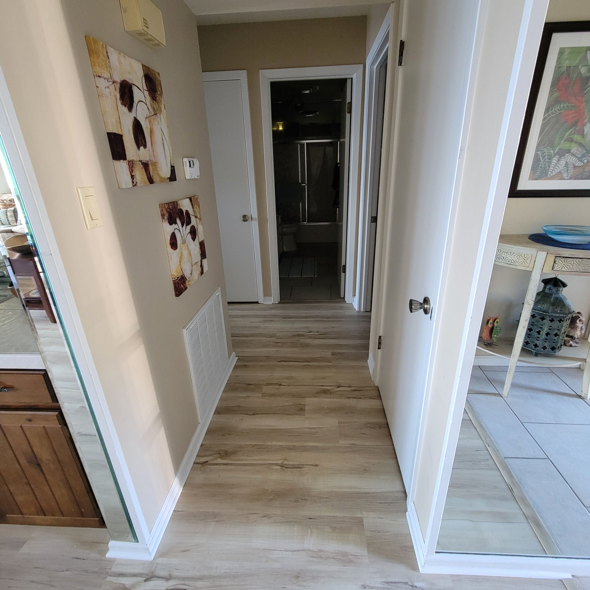 Narrow hallway with wood-look flooring, leading to a bathroom. Light beige walls, artwork, and white trim are present.