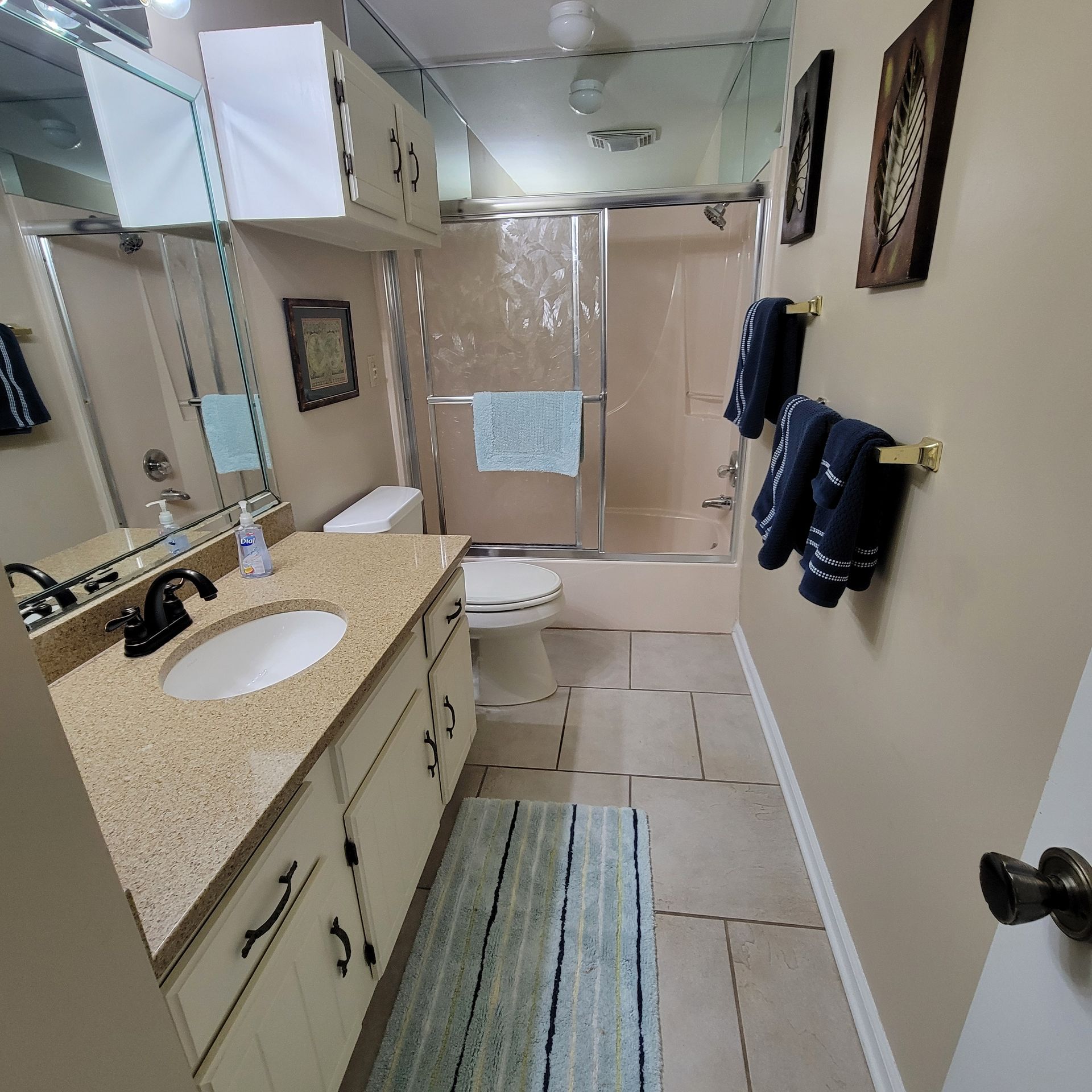 Bathroom with beige walls, vanity, toilet, and shower. Blue towels and a striped rug.