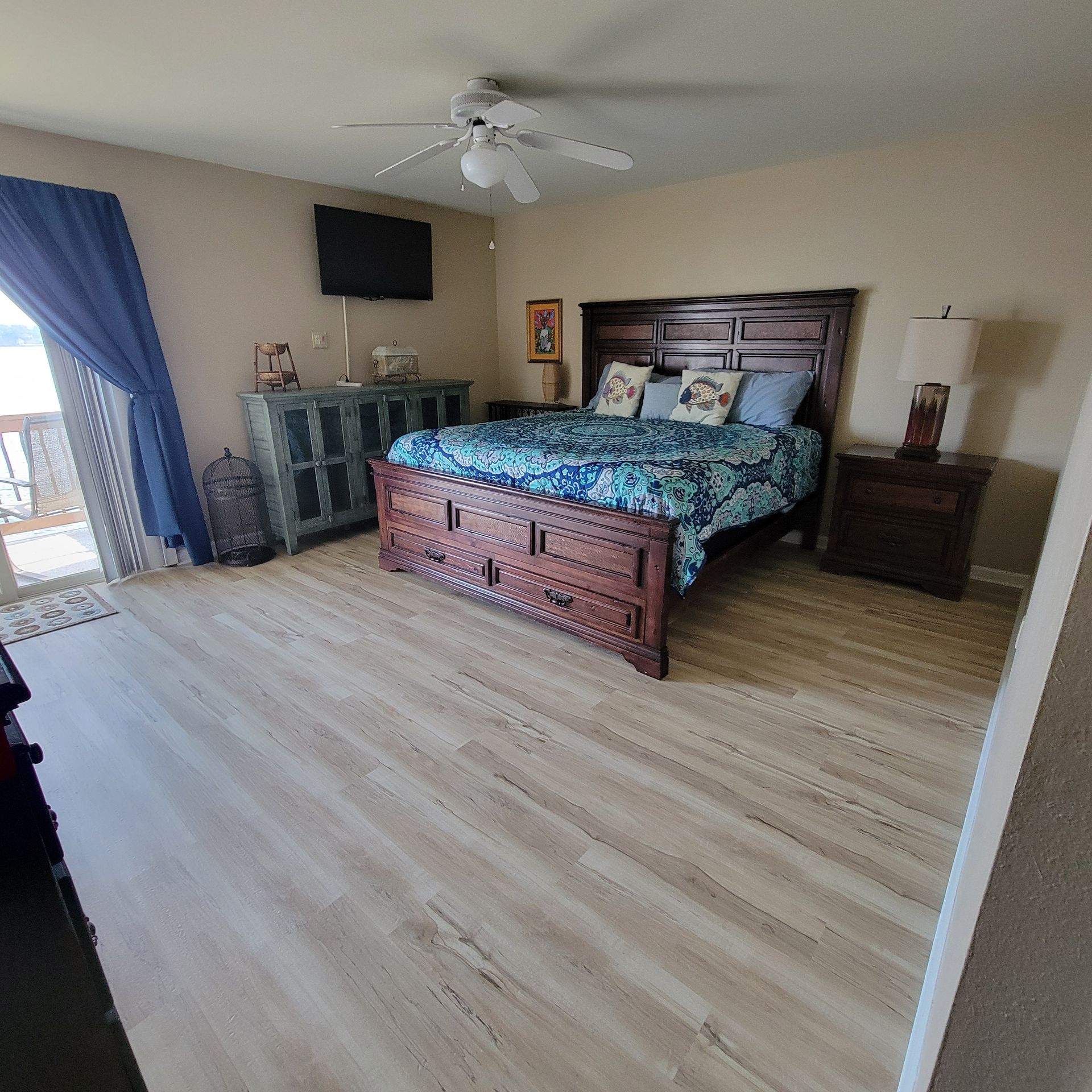 Bedroom with bed, dresser, ceiling fan, TV, blue curtains, and wood-look flooring.