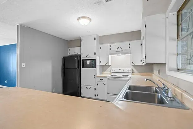A kitchen with white cabinets , a black refrigerator , a sink , and a window.