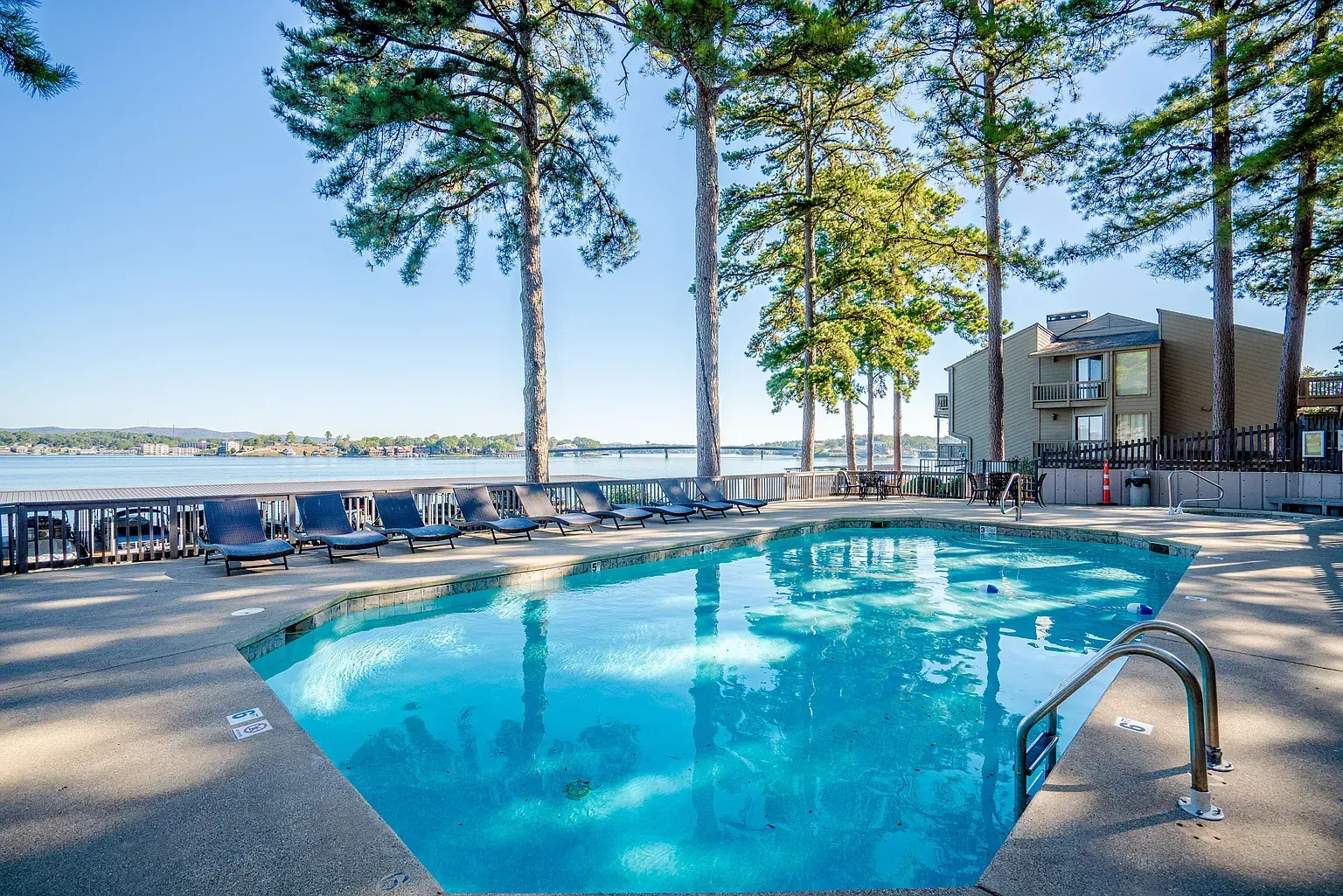 Swimming pool with lake view, surrounded by trees and lounge chairs. Sunny day, clear blue water.
