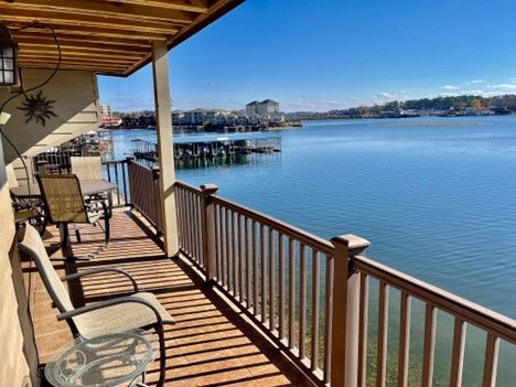Balcony overlooking a serene lake. Brown railing, table, chairs, and clear blue sky.