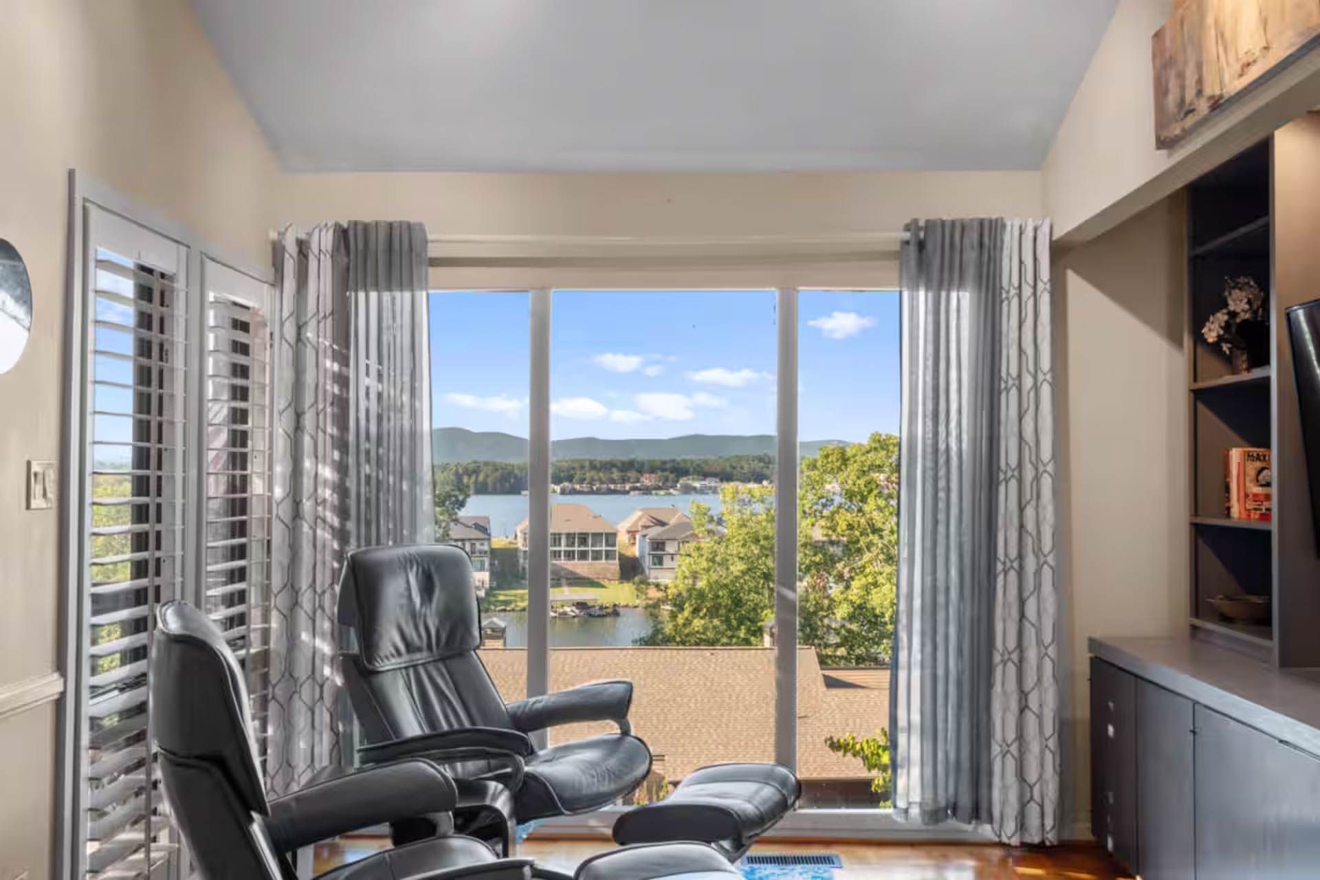 Two black recliners face large window with lake view; room with curtains, shelves, and shutters.