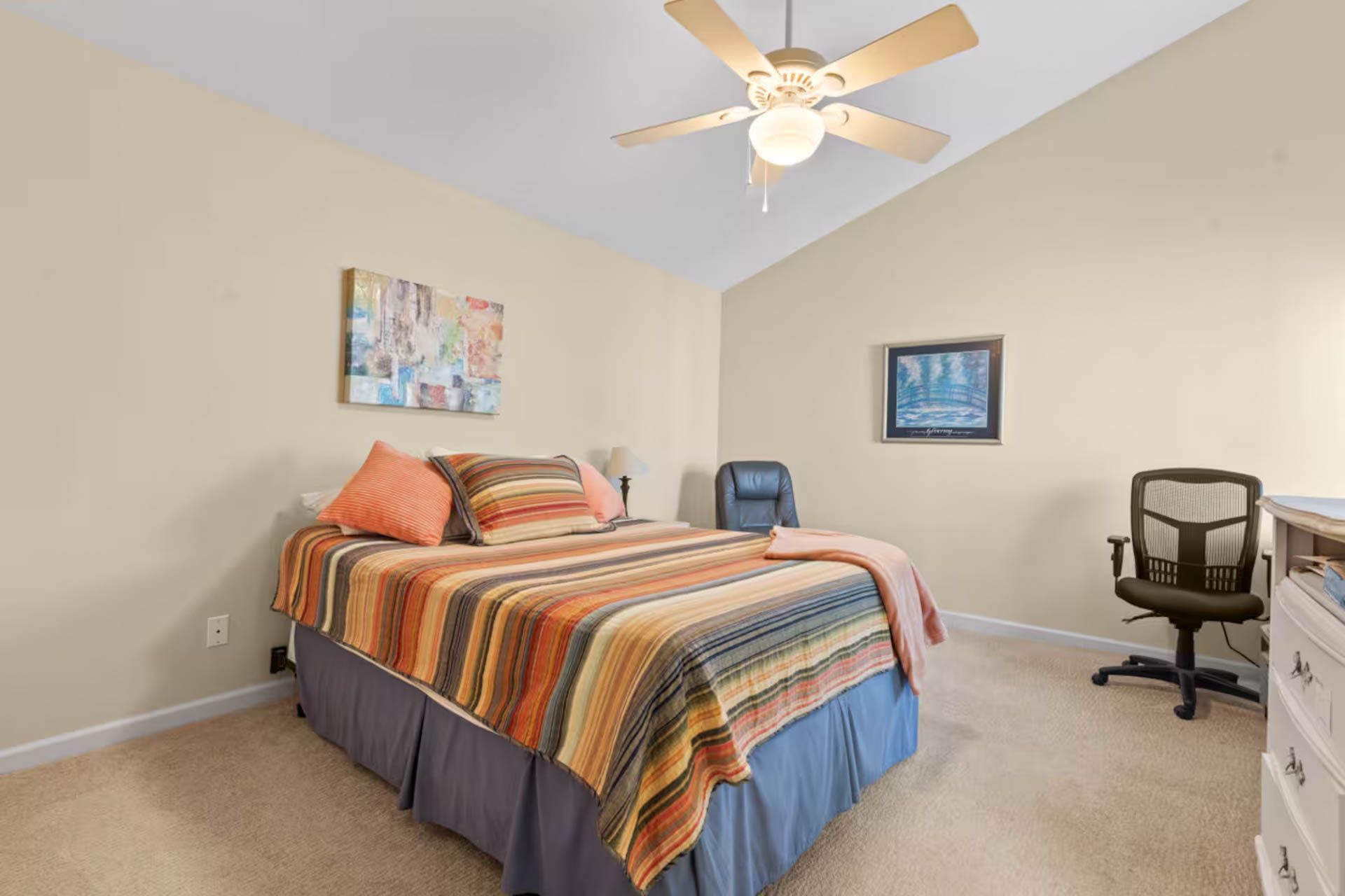 Bedroom with a bed, desk, chair, and art on beige walls; ceiling fan.
