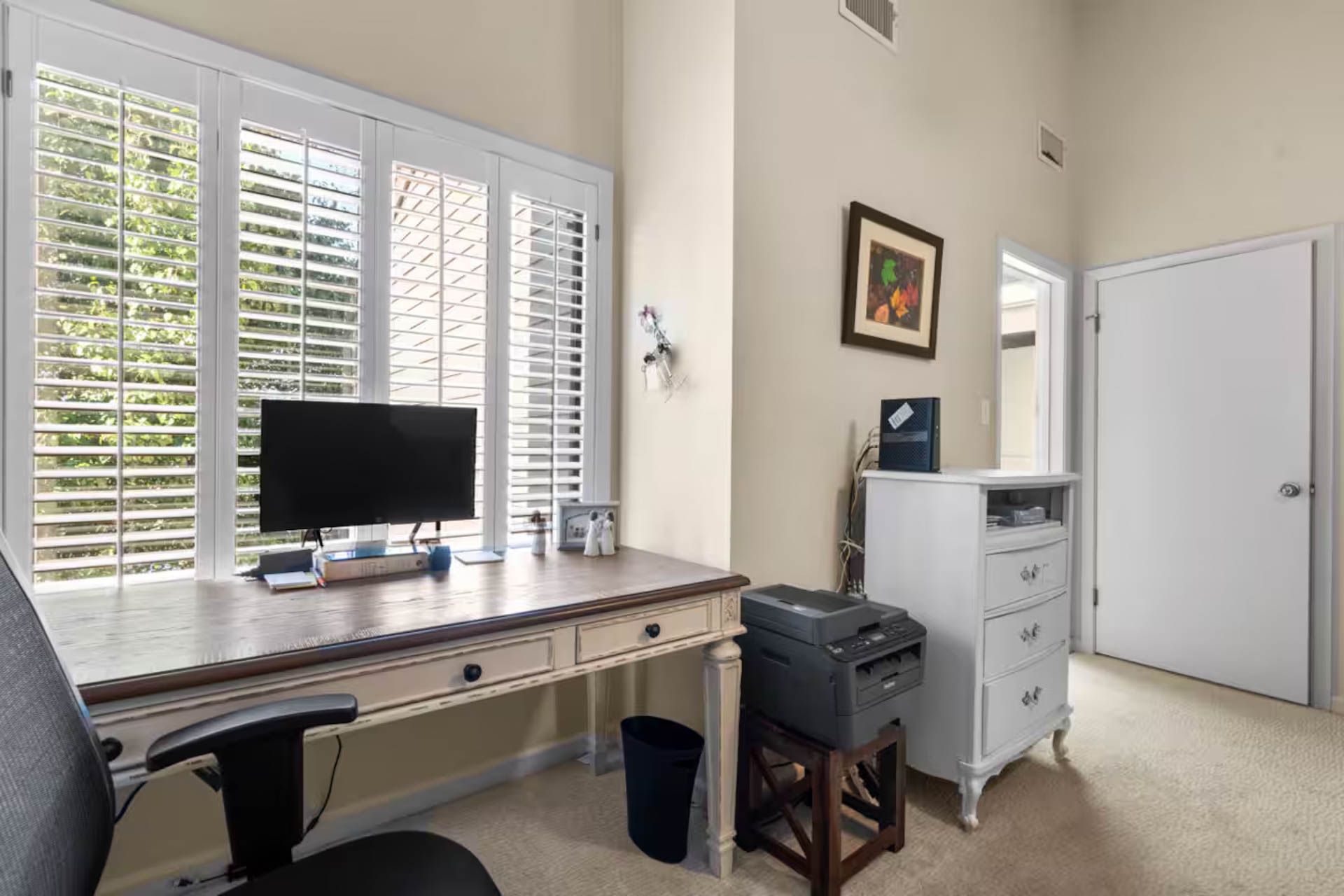 Home office with desk, window, computer, and printer. White shutters and cabinets, beige walls and carpet.