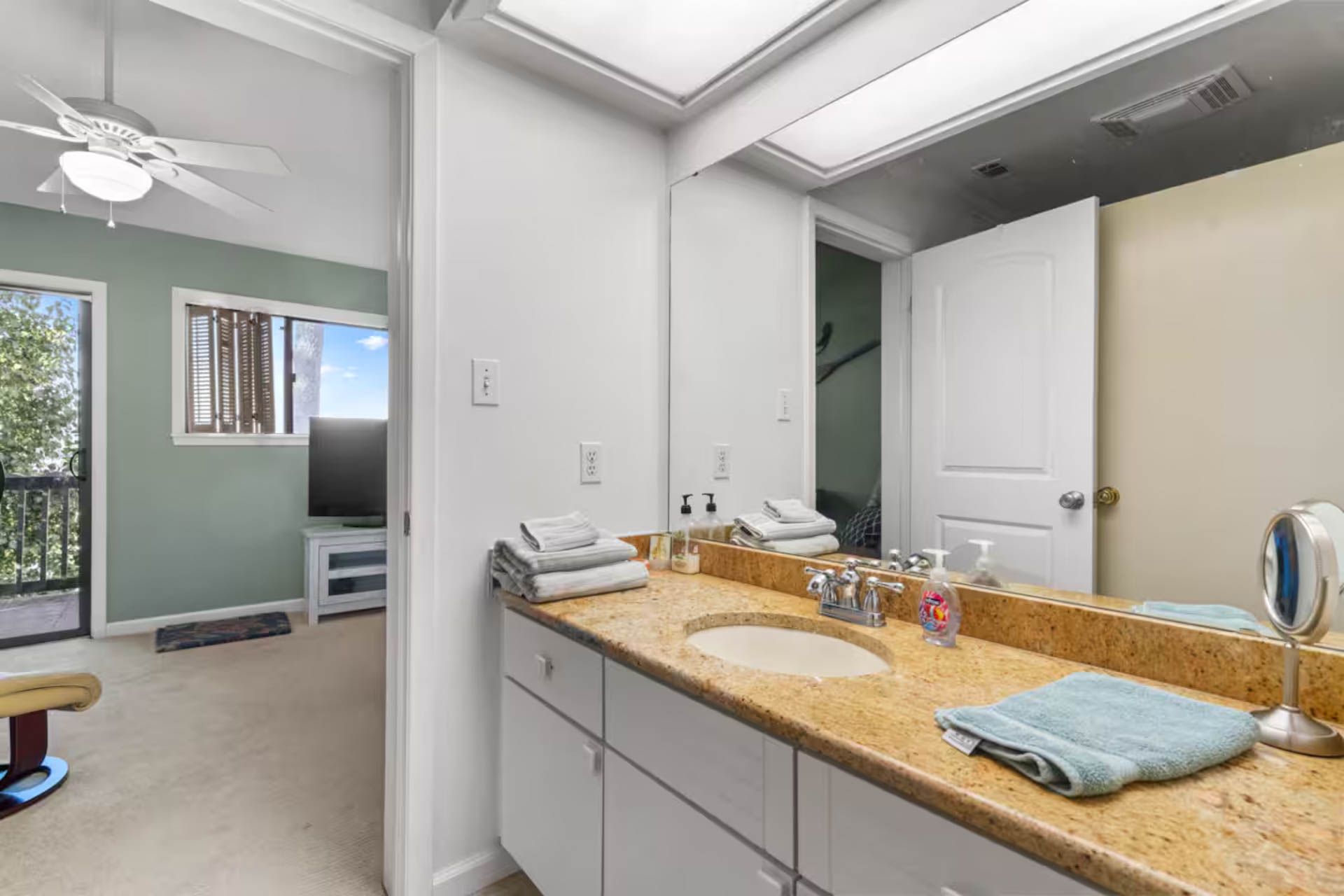 Bathroom with vanity, sink, and towels. Open door to living area with a TV and window.