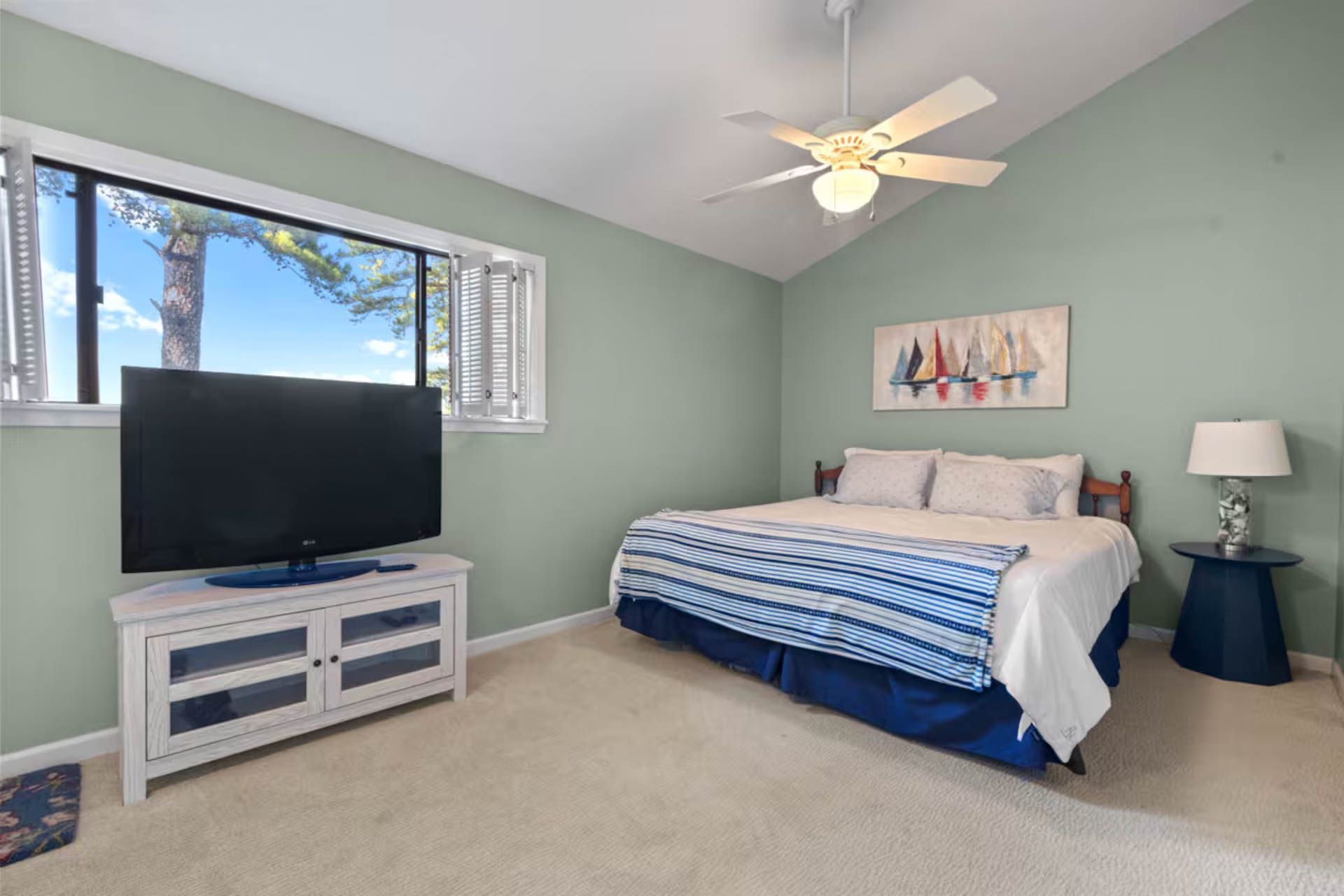 Bedroom with bed, TV, window overlooking a bright blue sky, light green walls, and beige carpet.