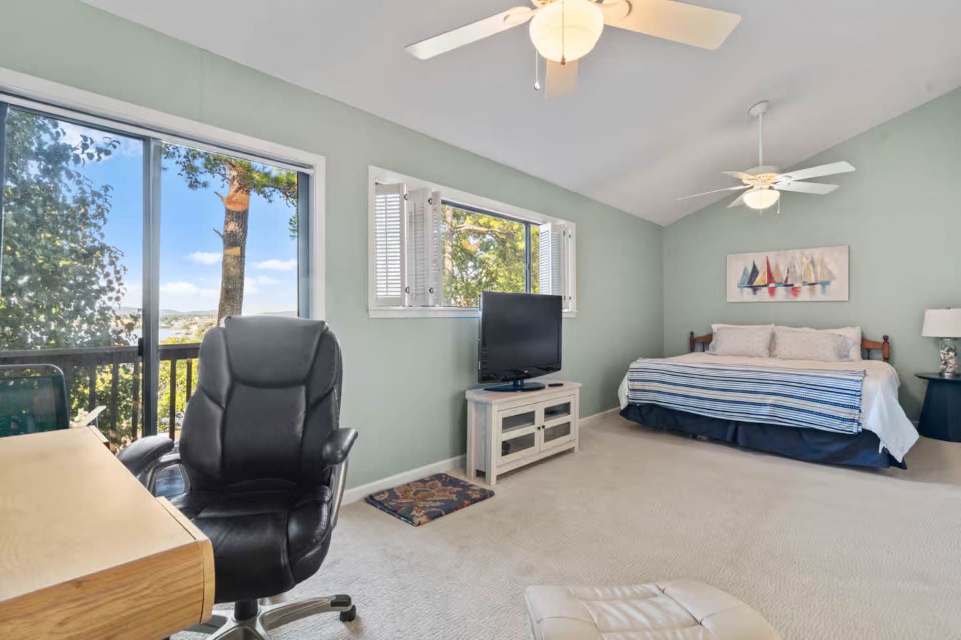 Bedroom with a desk, bed, TV, and large window overlooking a view. Light blue walls, white ceiling fans, and neutral carpet.