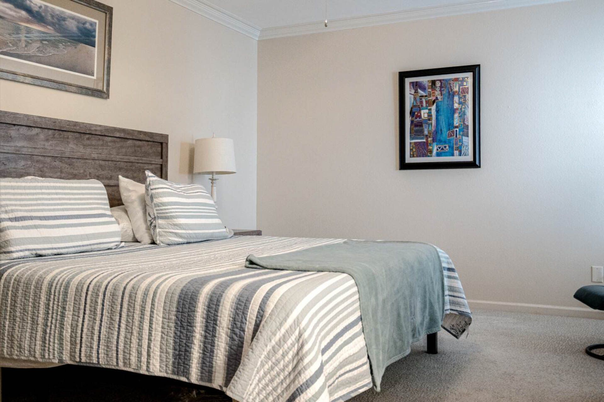 Bedroom with bed, striped quilt, pillows, lamp, and artwork. Neutral walls and carpet.