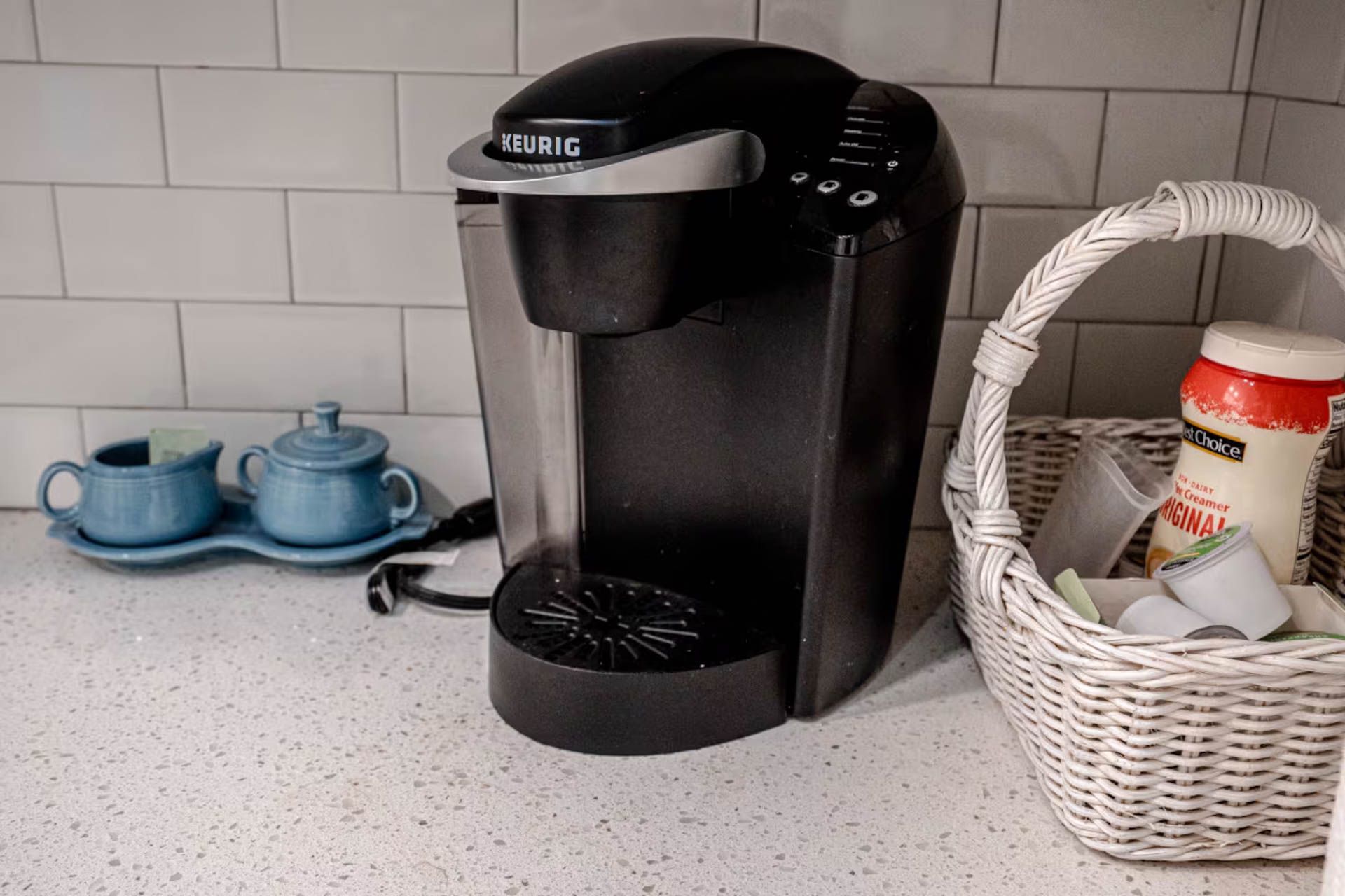 Black Keurig coffee maker on a countertop, next to a basket with coffee pods, near blue sugar bowls.