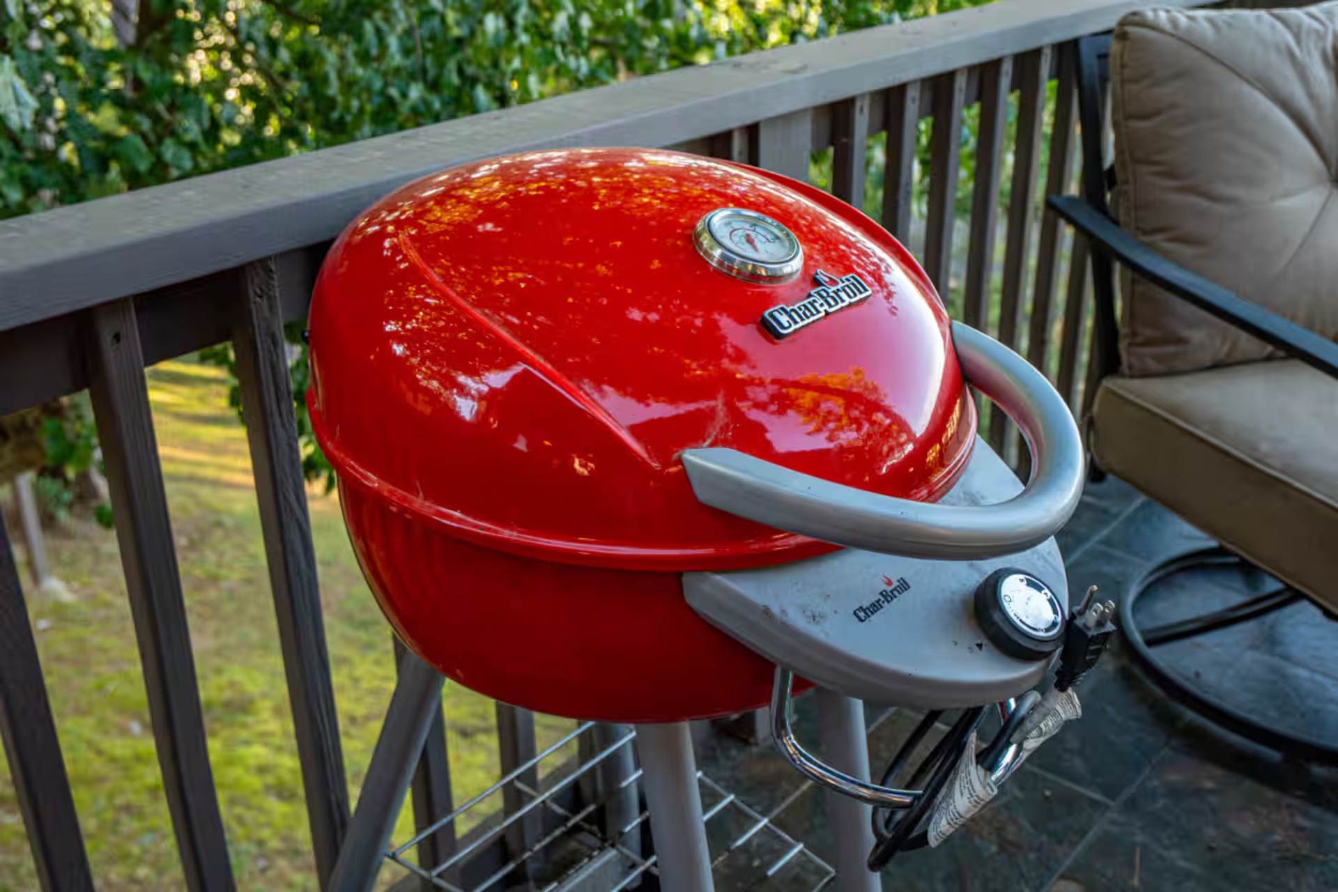 Red Char-Broil portable grill on a wooden deck with a chair, green foliage in background.