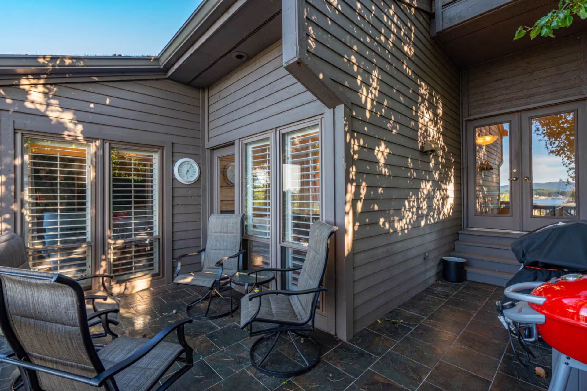 Patio with seating, shuttered windows, and a grill, against a brown house exterior.