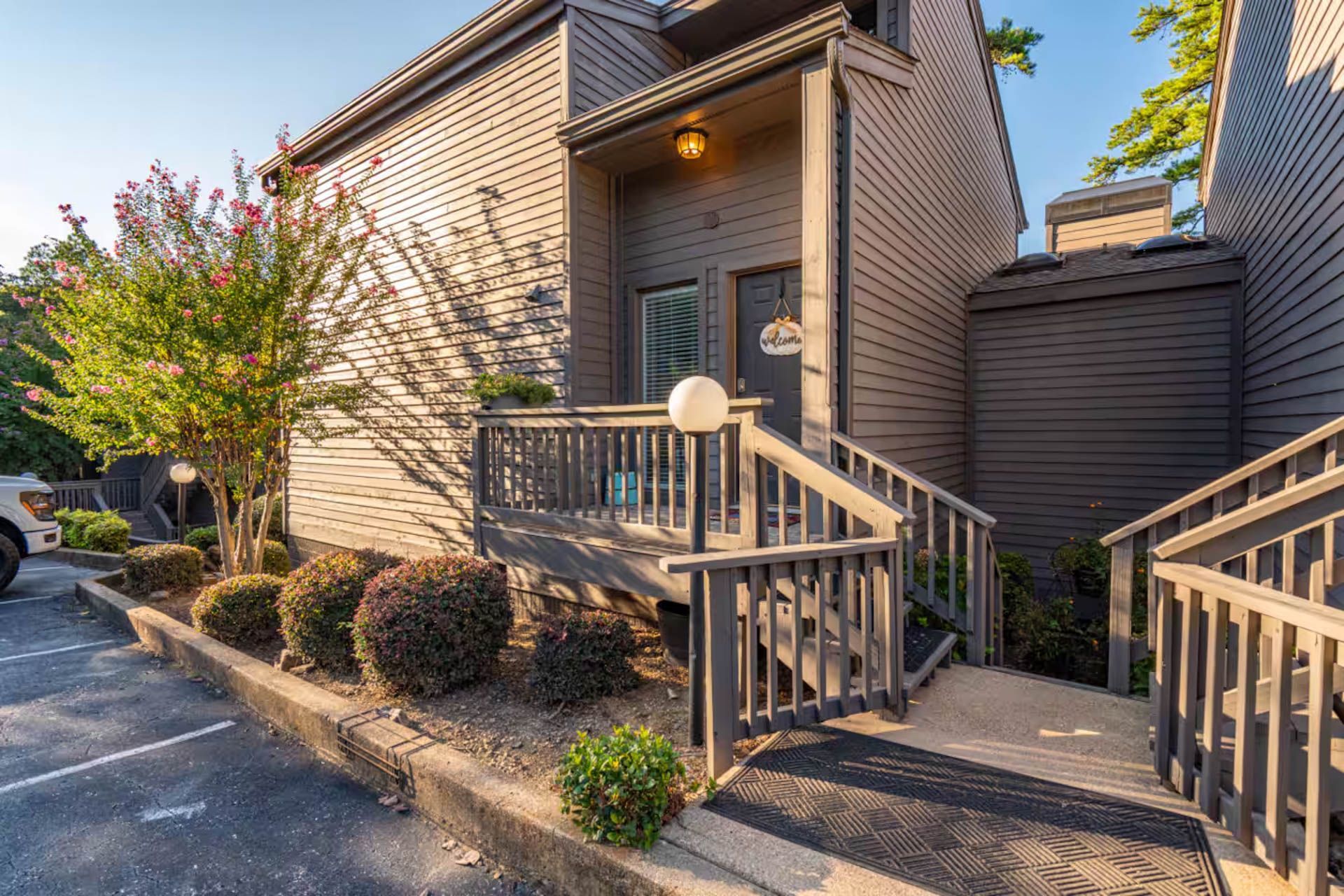 Gray townhouse with stairs and deck, shrubs, and parking space.