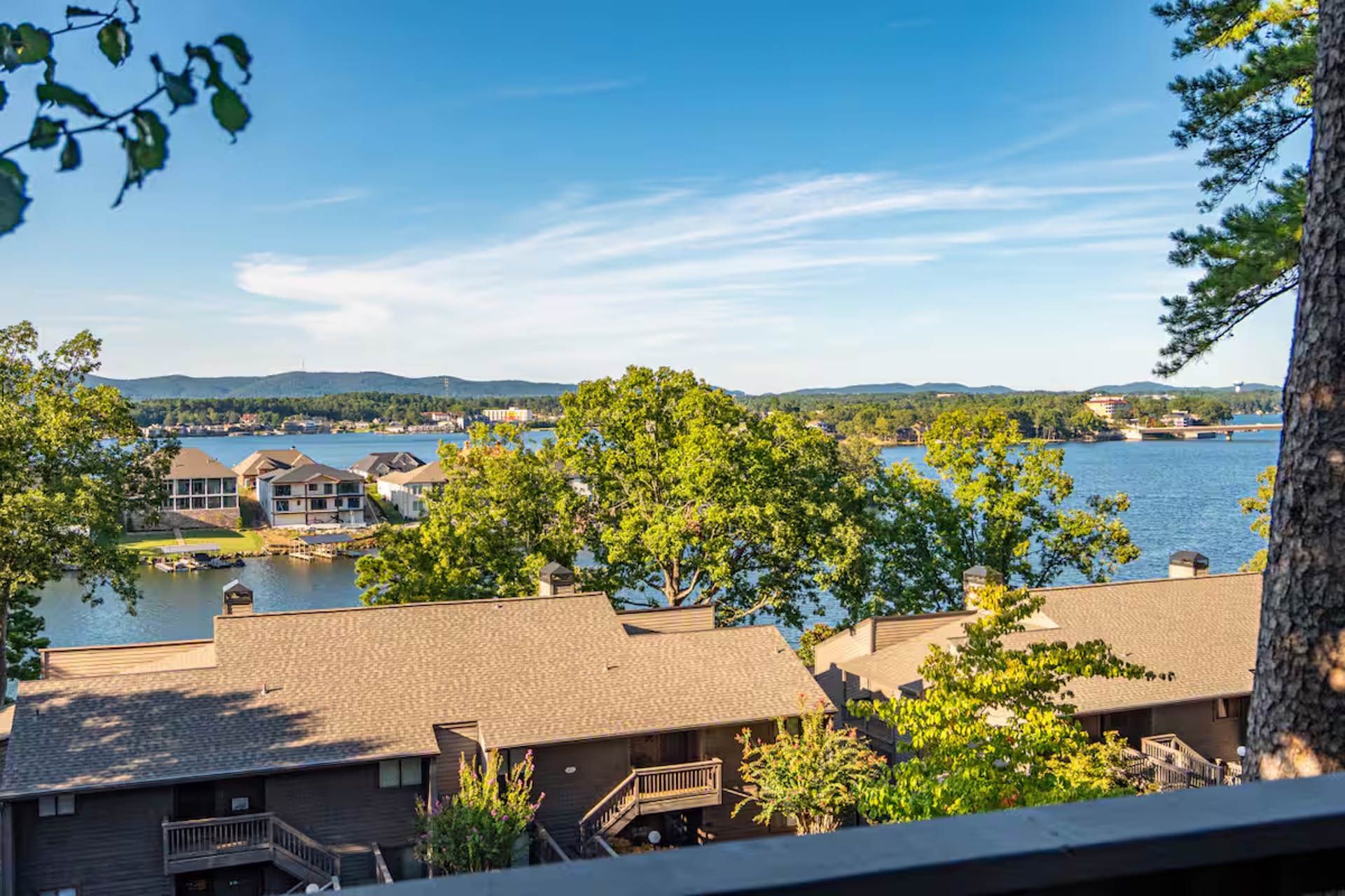 View of a lake and houses under a blue sky, seen from a slightly elevated position, trees in foreground.