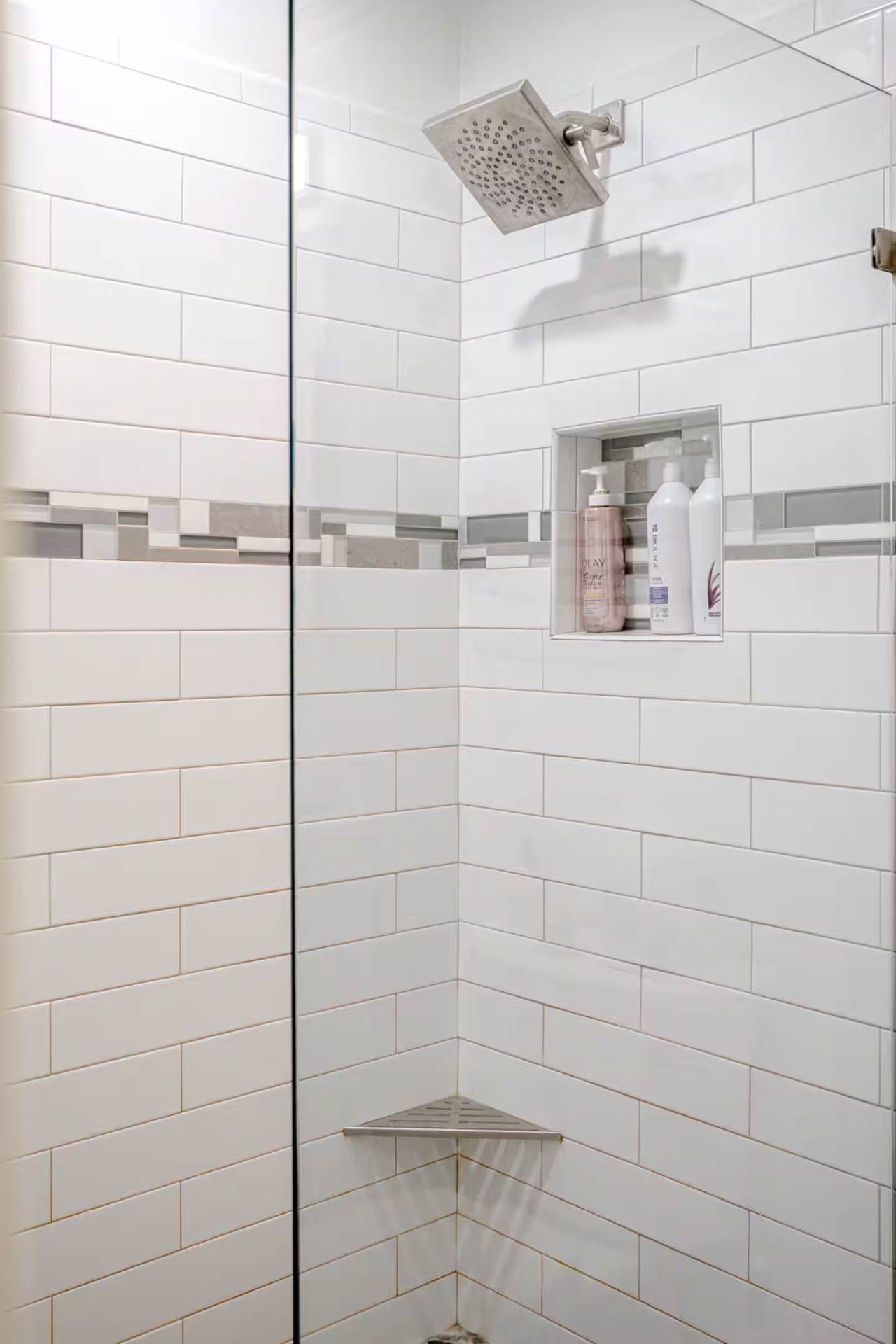 White-tiled shower with glass door and built-in shelf holding toiletries. Silver shower head and corner shelf.