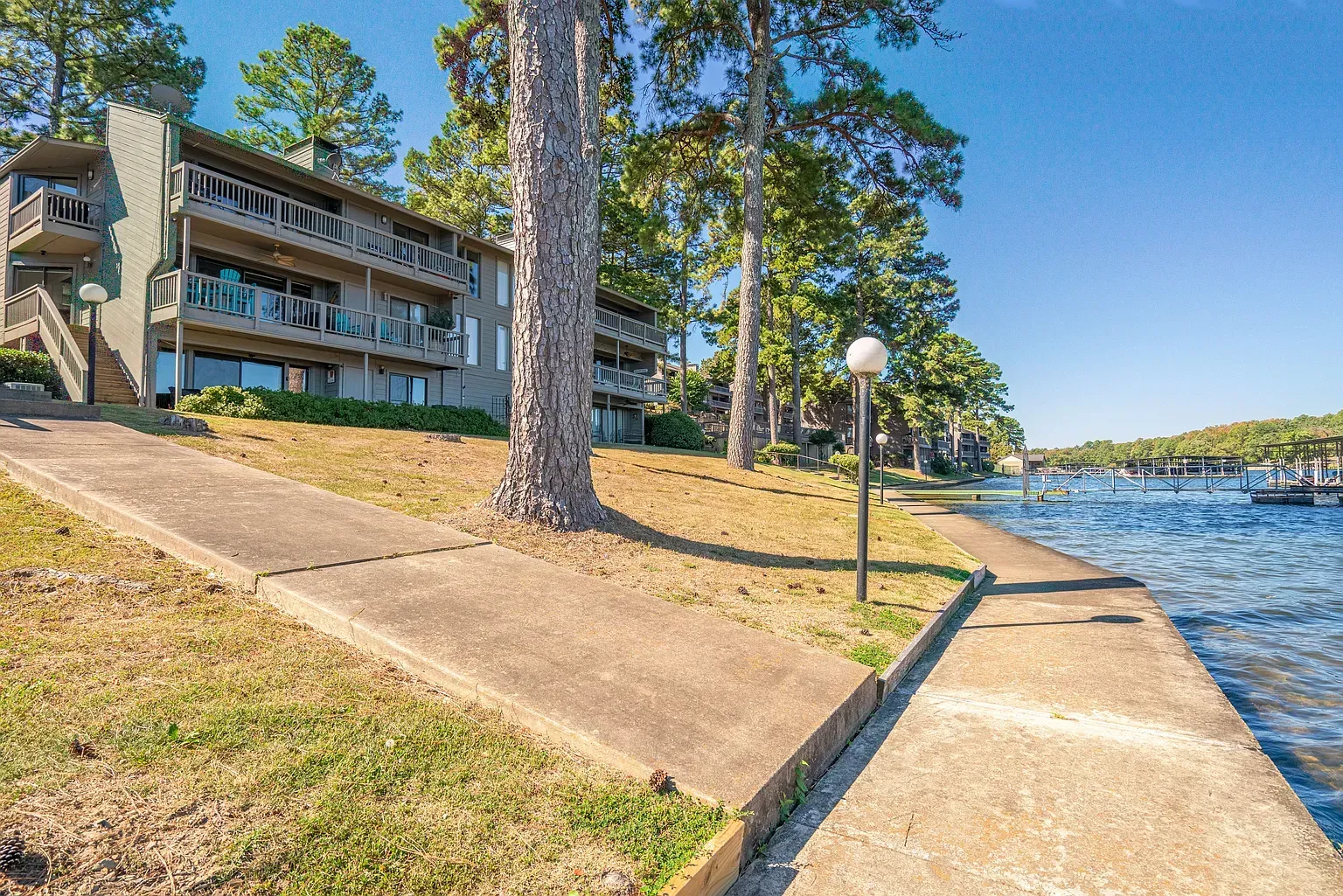 Gray building with balconies on a lake. Concrete path along the water with trees and blue sky.