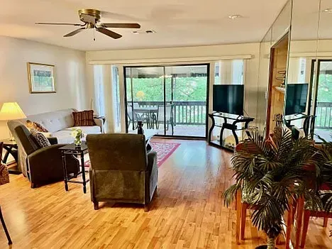 Living room with wooden floors, sliding door to balcony, sofas, chairs, TV, and ceiling fan.