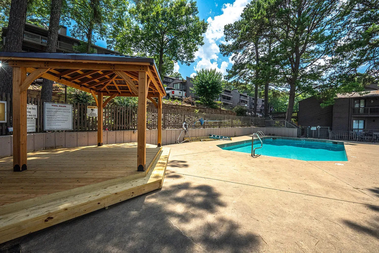 Wooden gazebo over a deck beside a turquoise pool in a sunny outdoor setting.