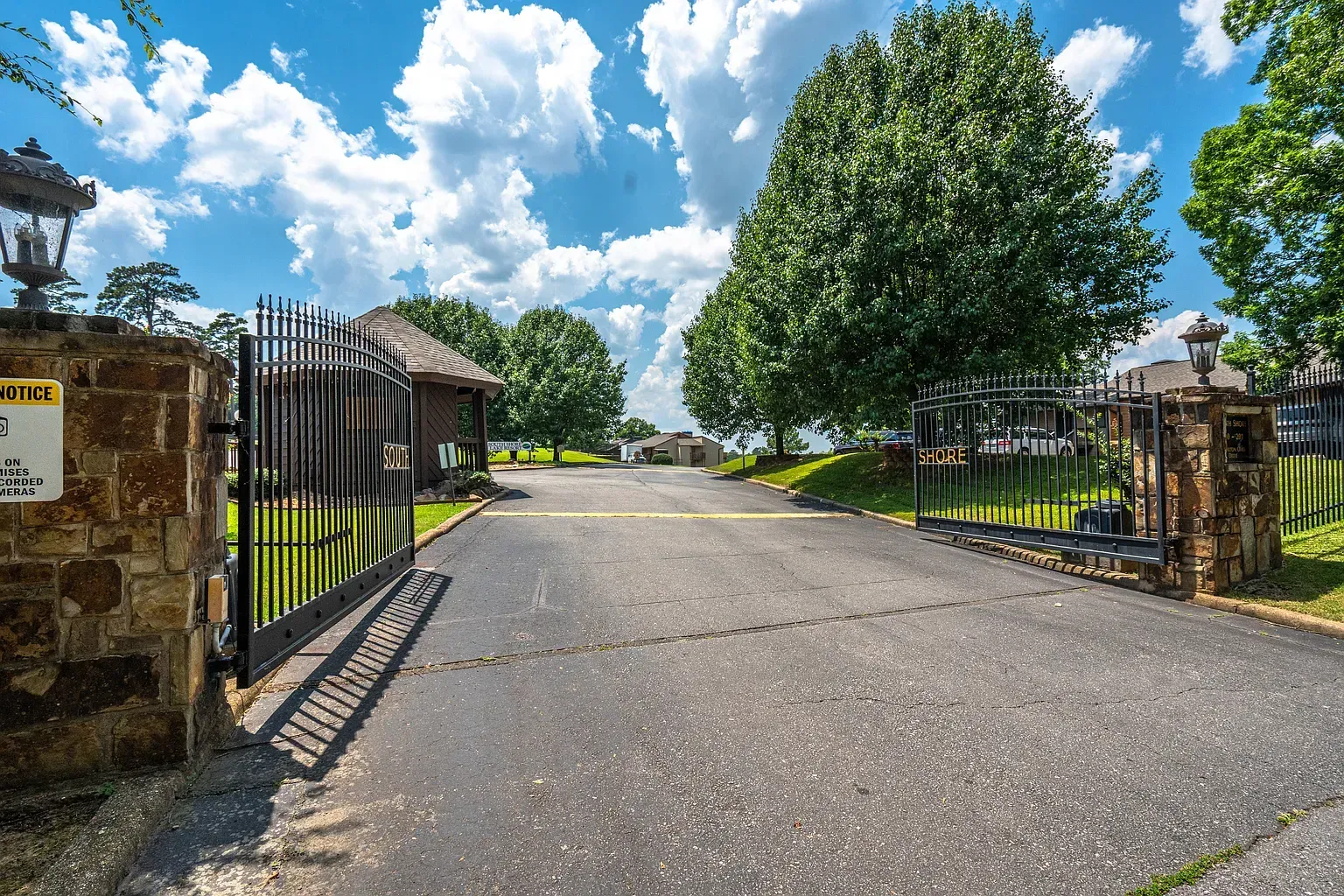 Gated entrance to a property with black iron gates, stone pillars, and a long asphalt driveway. Blue sky with clouds.