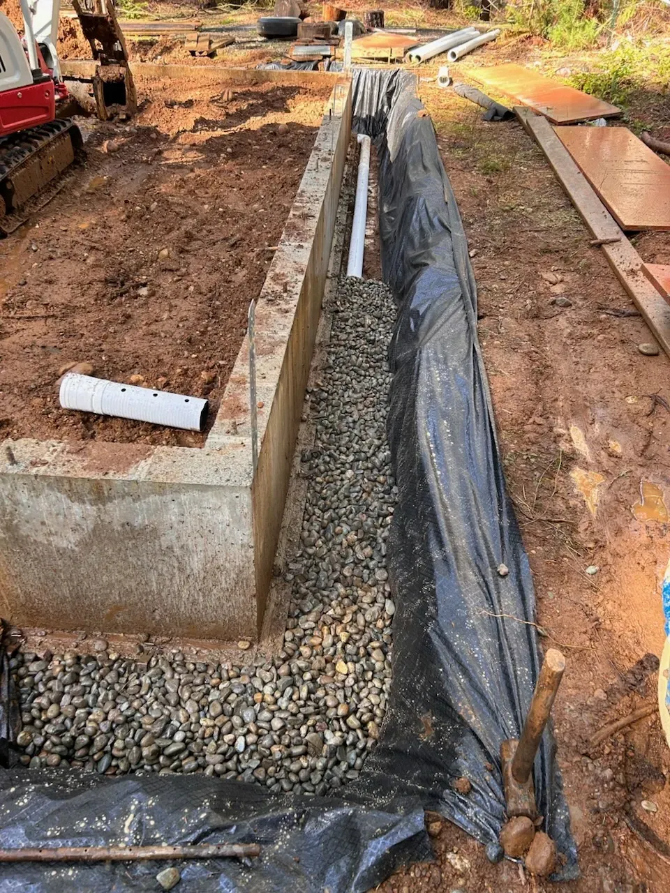 Construction trench lined with black plastic beside a concrete foundation, with gravel and pipes on site