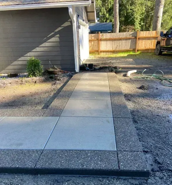 Concrete walkway leading to a gray house’s side entrance, with gravel yard and wooden fence in back.
