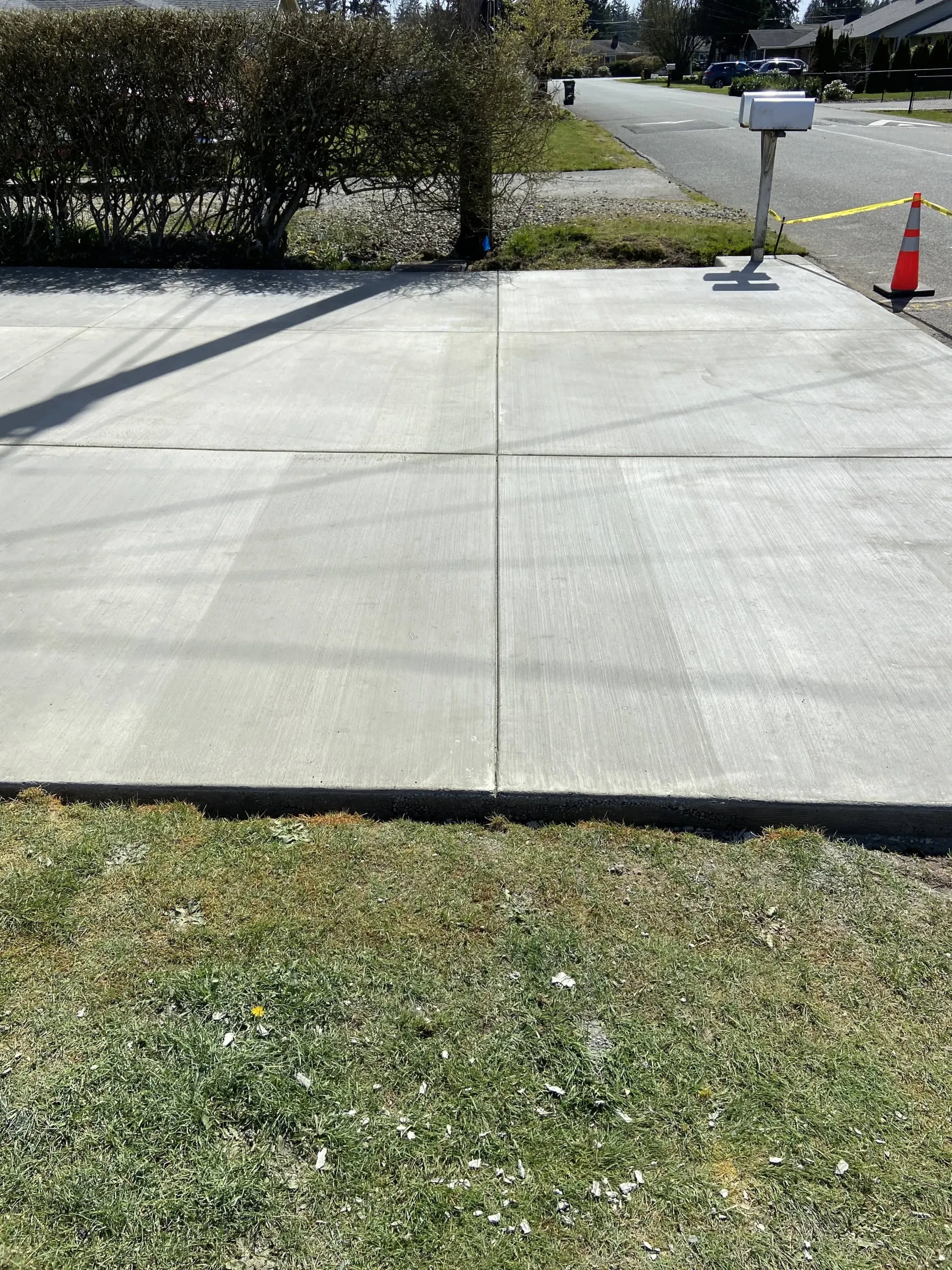 Concrete driveway leading to a street, with hedge, mailbox, and orange traffic cone on the right.