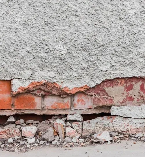 Cracked exterior wall with peeling gray stucco exposing red bricks and rubble at the base
