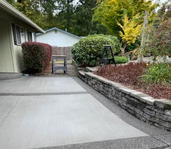 A light-colored concrete patio leads to a stone retaining wall, bushes, and a partial view of a house in a garden setting.
