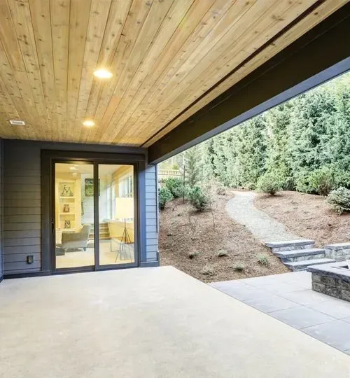 Covered patio with recessed lights, glass doors, and steps leading to a wooded hillside path.