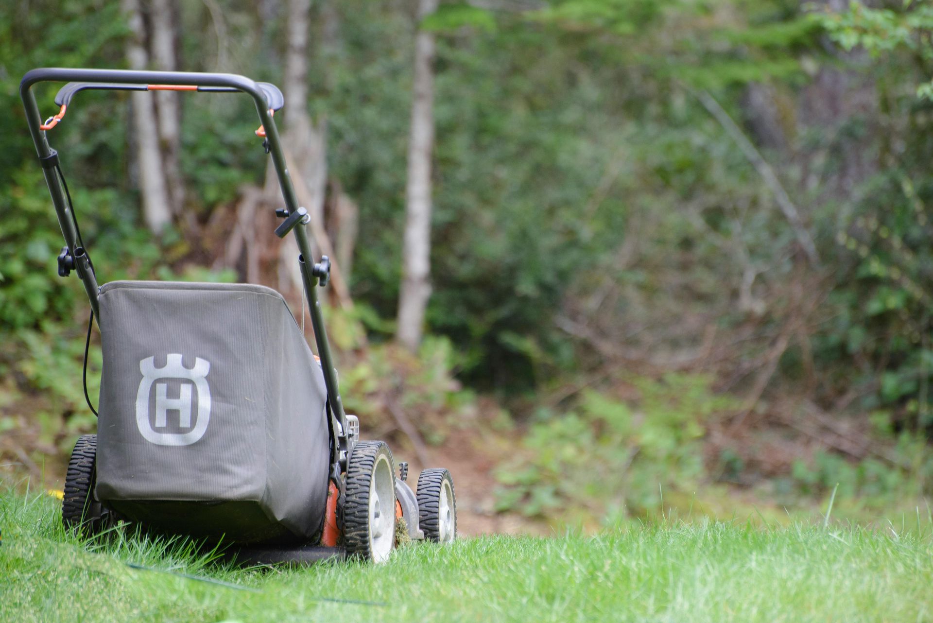 Gray lawn mower on grass with a black handle and Husqvarna logo, set against a blurred wooded background