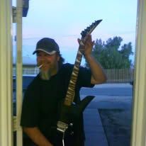 Man holding a black electric guitar, smoking, standing in a doorway at dusk.