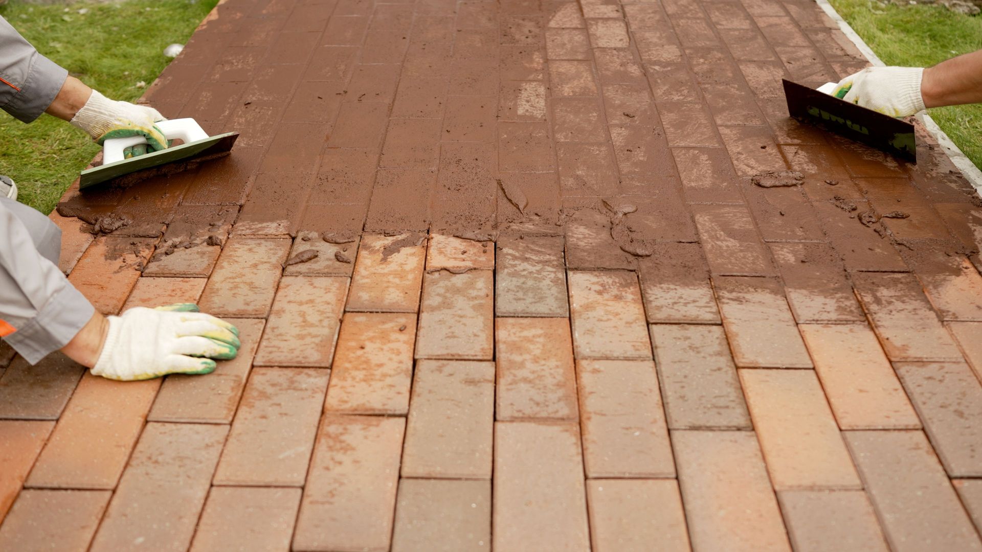 Two people in work gloves use trowels to spread dark brown mortar over a newly laid brick walkway.