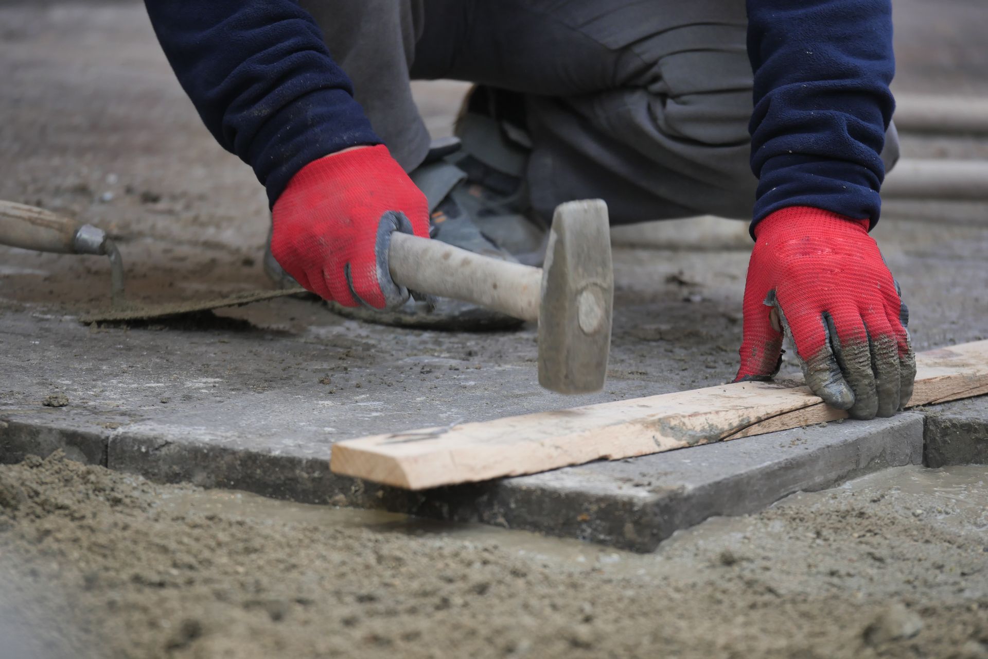 A worker wearing red gloves uses a hammer to tap a wooden board on top of newly laid stone pavers. A worker wearing red gloves uses a hammer to tap a wooden board on top of newly laid stone pavers.