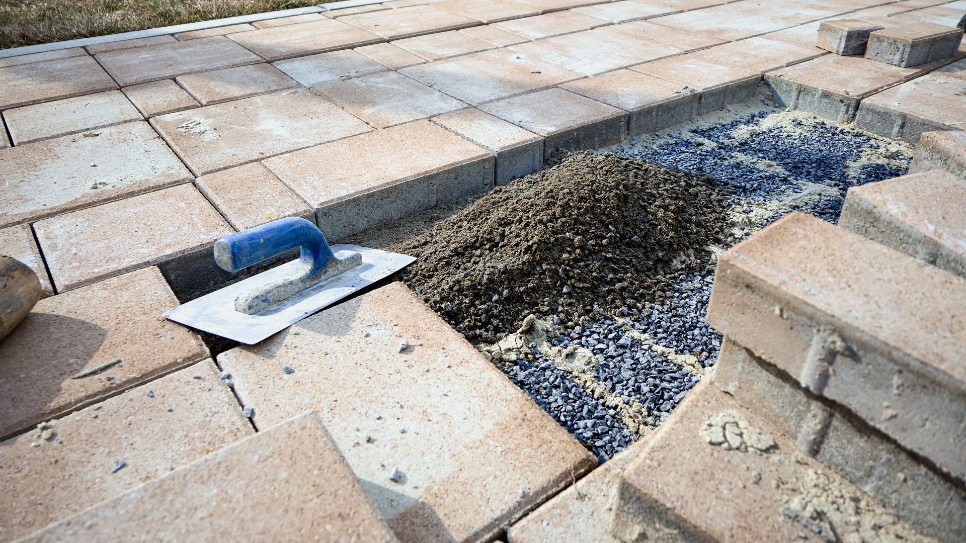 A metal trowel sits on tan paving stones next to a small section of unfinished, gravel-filled path under construction. A metal trowel sits on tan paving stones next to a small section of unfinished, gravel-filled path under construction.