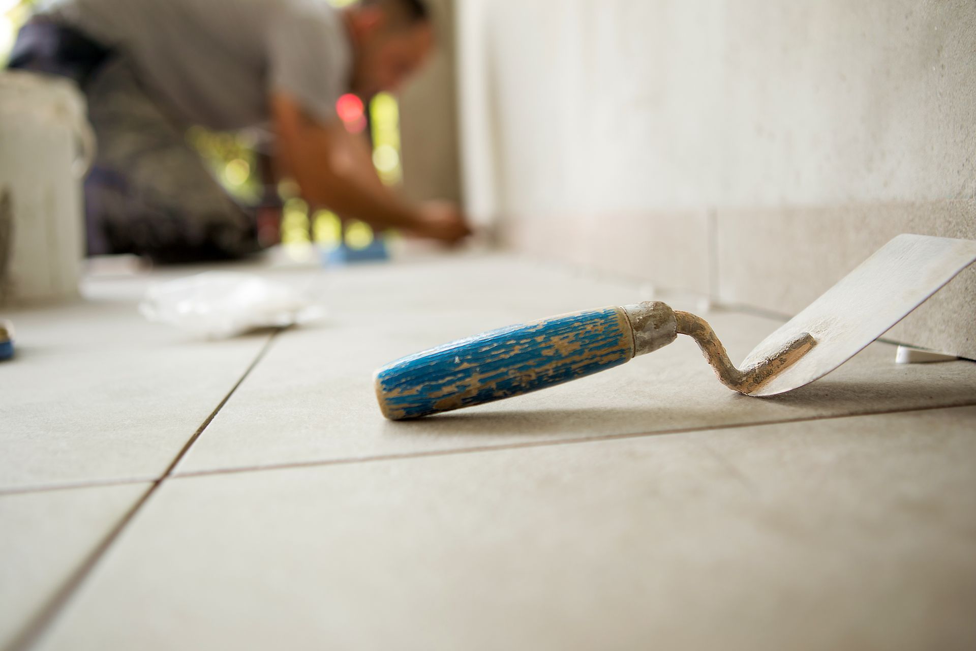 A close-up of a blue-handled trowel resting on newly laid light-colored tiles, with a person working in the background. A close-up of a blue-handled trowel resting on newly laid light-colored tiles, with a person working in the background.