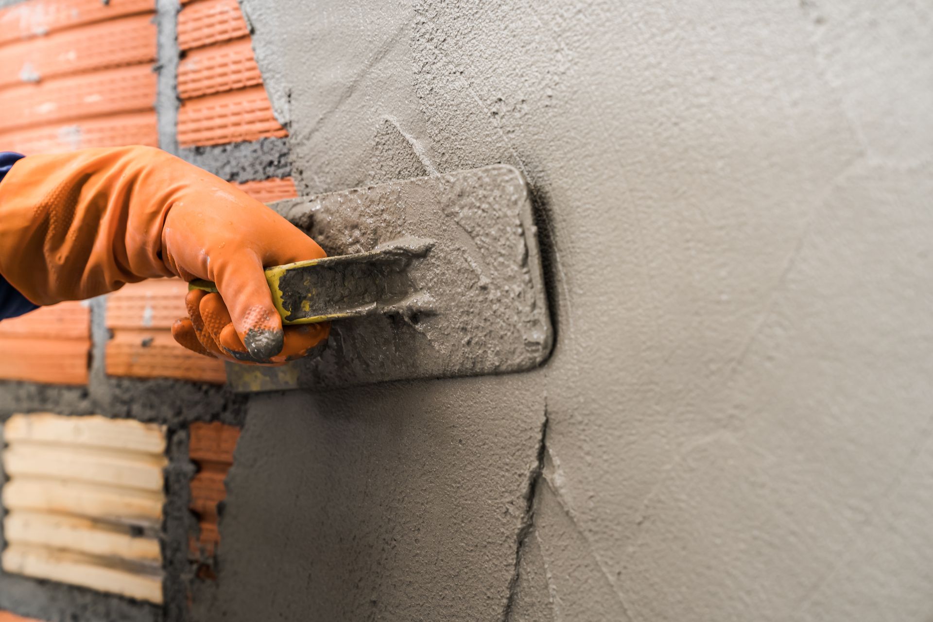 A hand in an orange glove uses a trowel to apply wet mortar over a red brick wall.