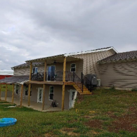 A multi-level home with tan siding, a wraparound wooden deck, and a lower-level patio, set on a grassy, sloping landscape.