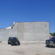 A black Jeep and a black truck parked in front of a large, blank, light-colored concrete wall under a clear blue sky.