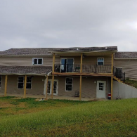 A two-story tan house with a covered patio on the ground level and a wooden deck balcony above it on a grassy hill.