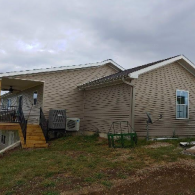 A tan vinyl-sided house with a wooden staircase leading to a covered porch under a cloudy sky.