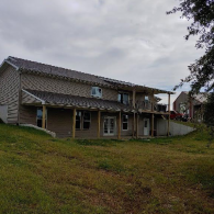 A two-story, tan-sided house with a metal roof and a covered porch, situated on a grassy, sloped lot under a cloudy sky.