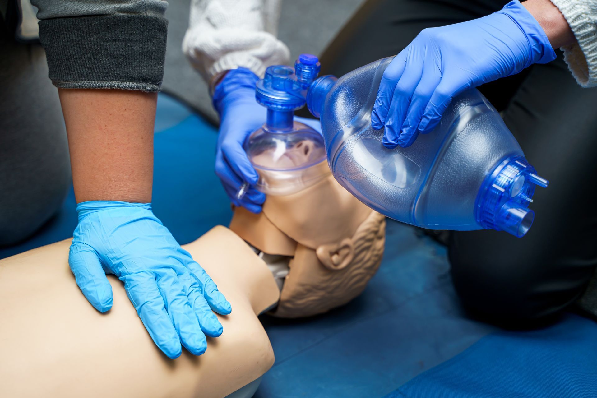 Two people in blue medical gloves perform CPR on a training mannequin using a bag valve mask.