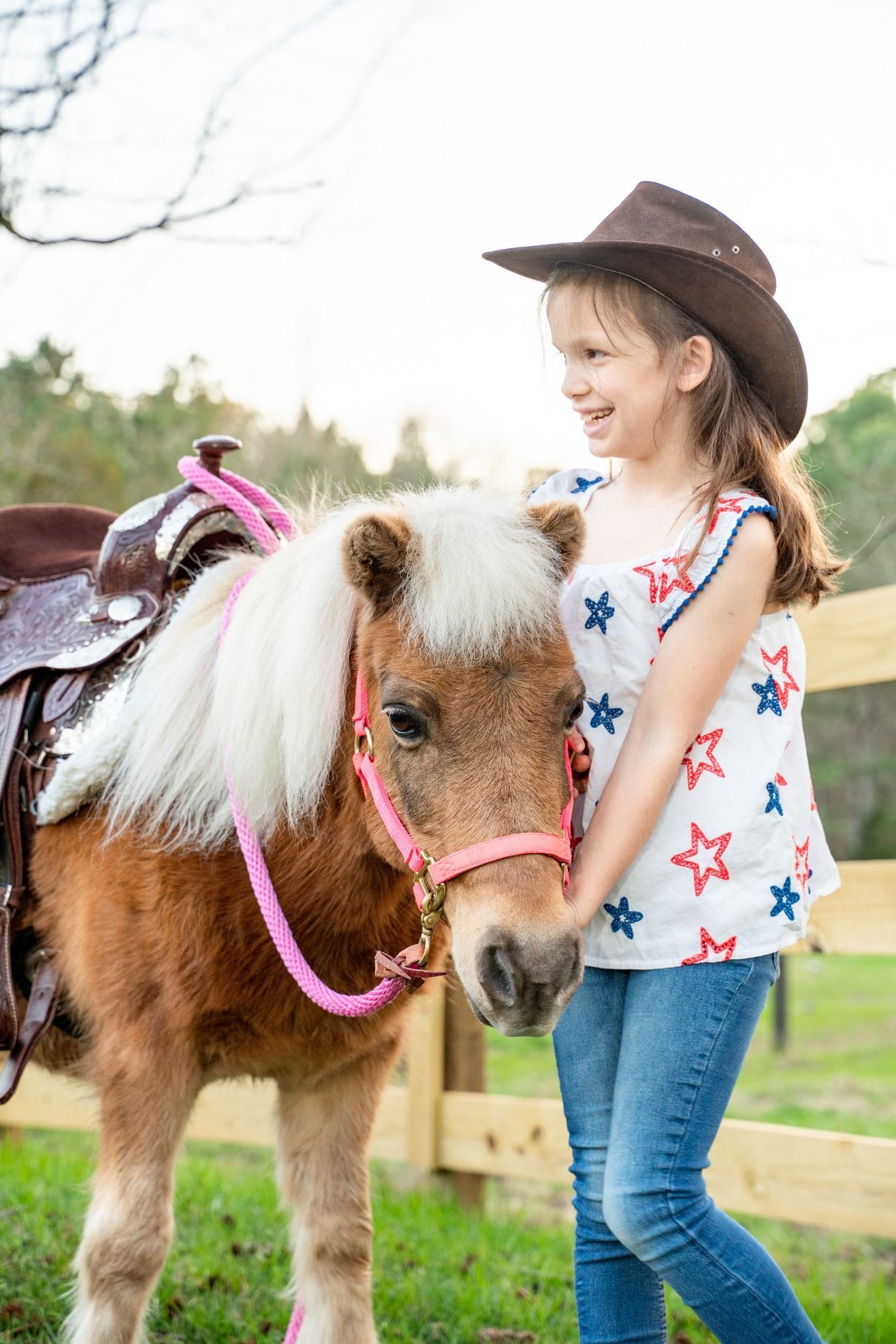 A little girl in a cowboy hat is standing next to a small pony.