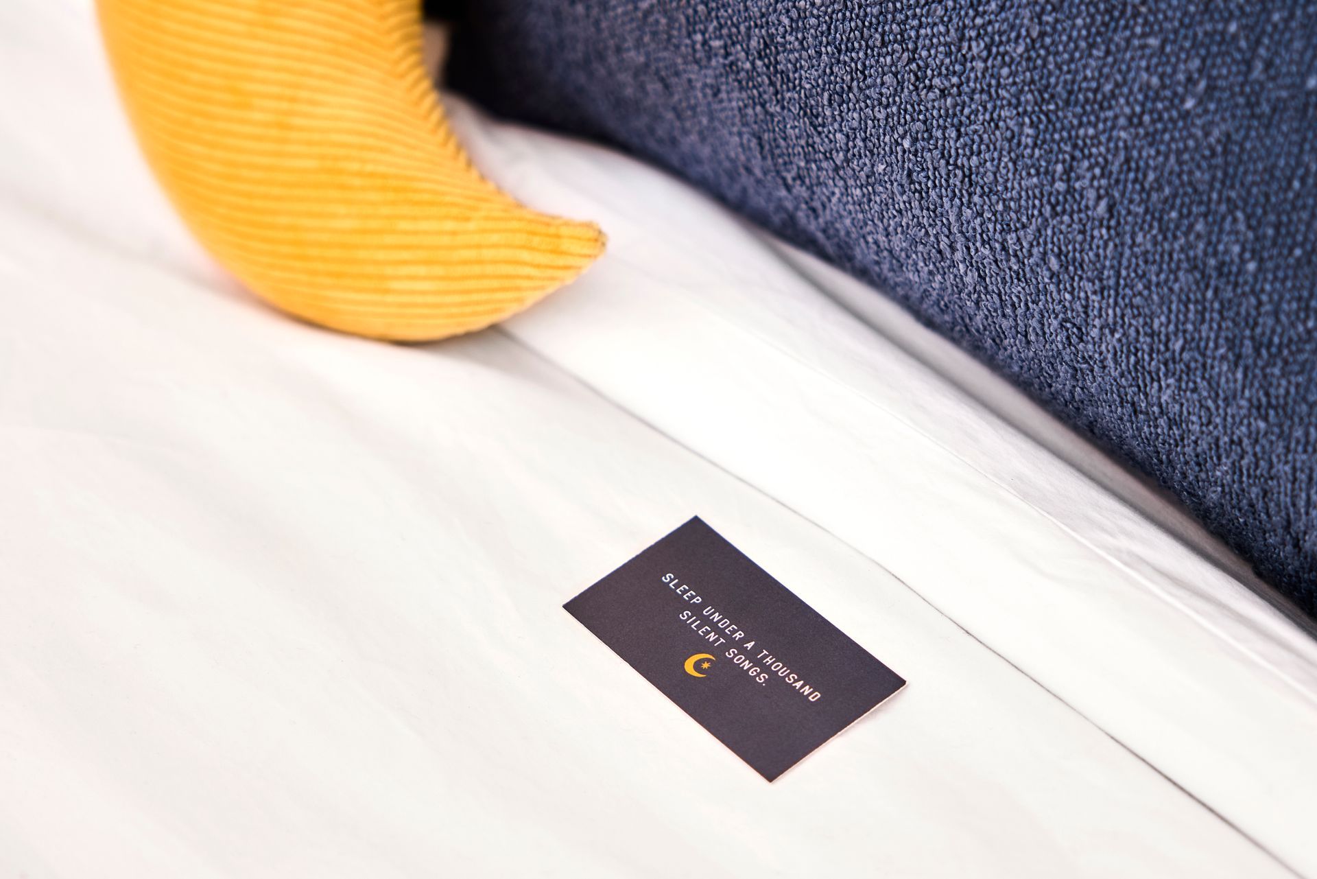 White bed with a dark card and orange pillow against a textured navy cushion