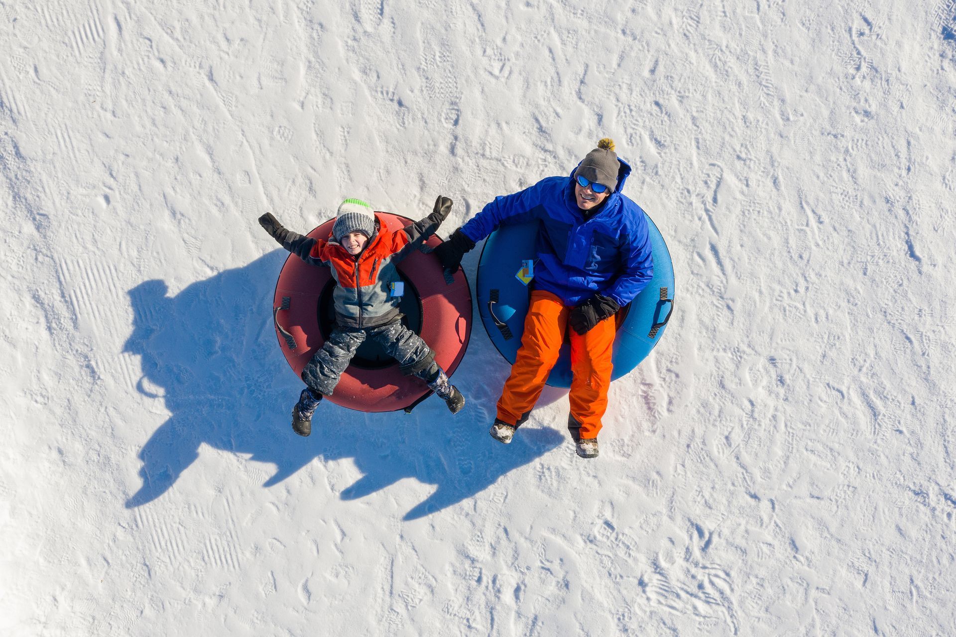 Two people tubing down a snowy hill. One in red, one in blue, enjoying the winter fun.
