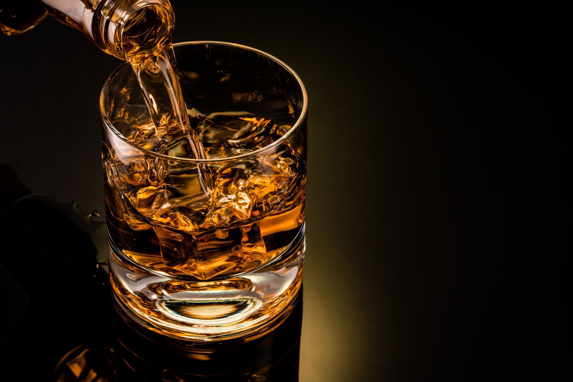Whiskey being poured into a rocks glass filled with ice, against a dark background.