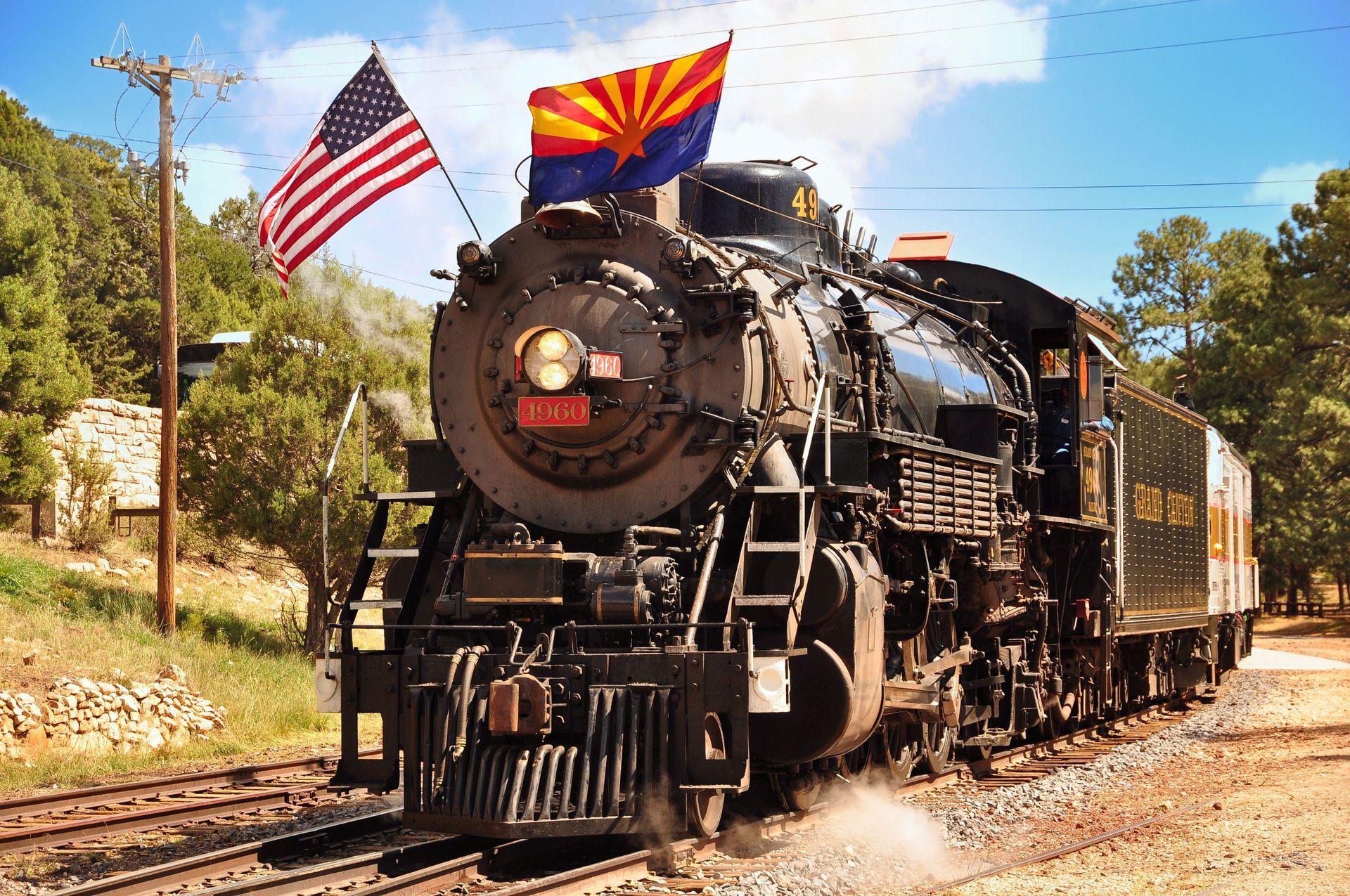 Steam train with US and Arizona flags on tracks in a sunny setting.